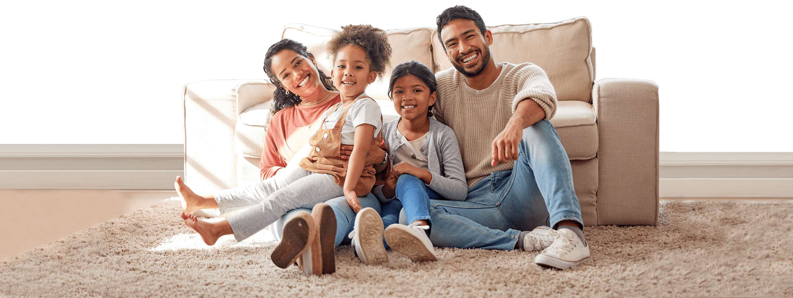 A family of four sits together on a soft beige carpet in a cozy living room. The parents, a man and a woman, smile warmly with their two young children between them. The room features a large, light-colored sofa.
