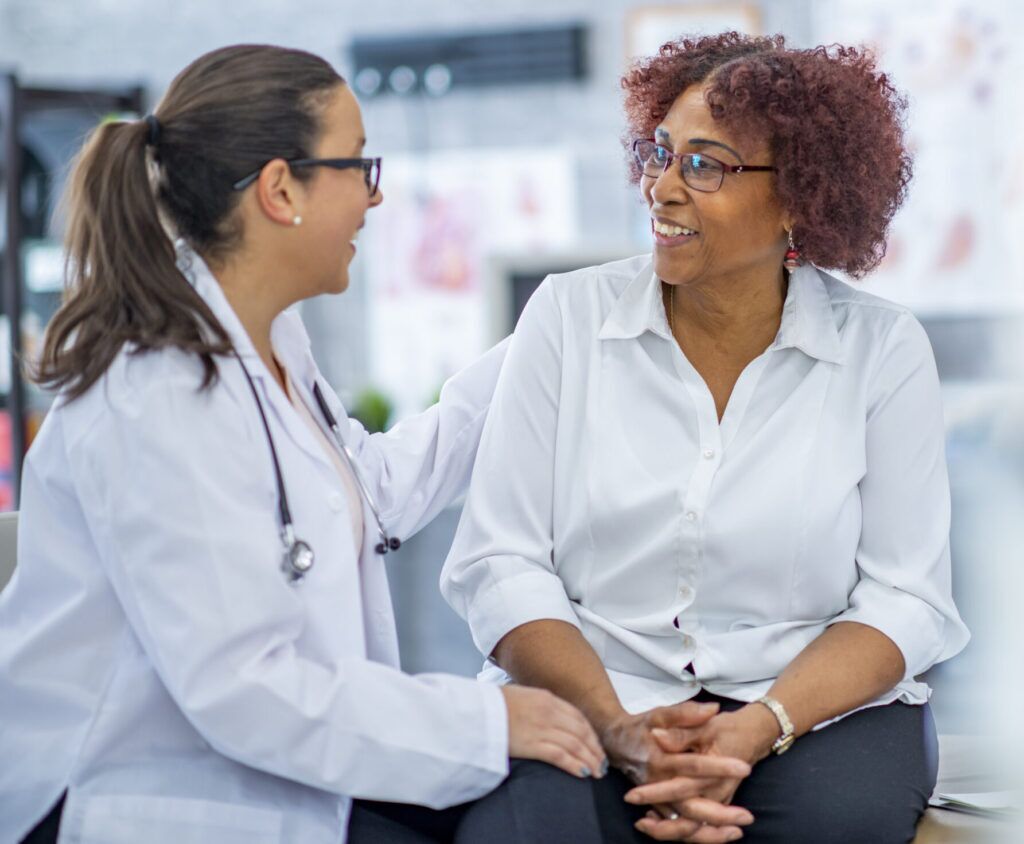 A healthcare professional with a stethoscope smiles and talks with an older woman sitting on an exam table. Both are wearing white shirts, and the setting appears to be a medical office.
